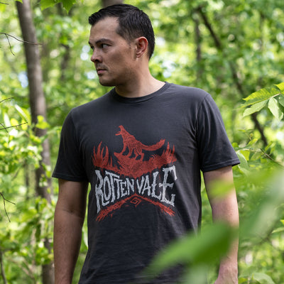 This image shows a photo of a man staring off at something in the forest. He is wearing the Monster Hunter Rotten Vale shirt and is looking very intent.
