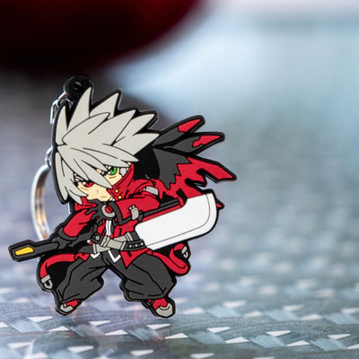 Image featuring the Ragna keychain, a piece of Blazblue merchandise, elegantly positioned on a glass surface with a beautifully blurred background.