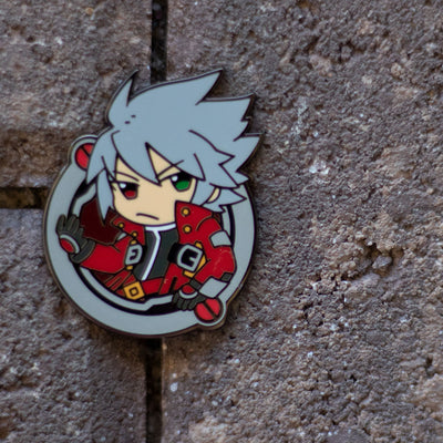 Image showcasing the Ragna pin, a piece of Blazblue merchandise, set against a textured brick background.