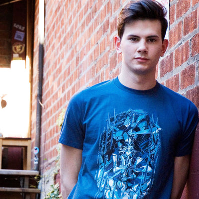 Photo of a young man wearing the Blazblue Frostbite shirt. He is looking directly at the camera, and is leaning against a brick wall.