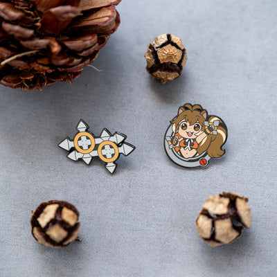 Both pins from the BlazBlue Makoto Pin Set elegantly arranged on a table, surrounded by pinecones for decoration.