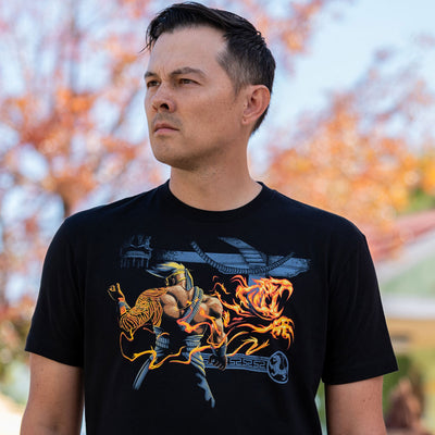 This photo is of a young man wearing the Killer Instinct Jago t-shirt. It shows him standing in front of a blurred out orchard of trees with yellow, orange and red leaves that are blurred out.