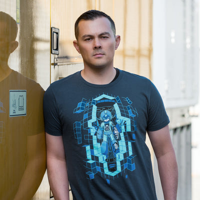 Image shows a young man wearing the Mega Man Upgrade shirt. He is leaning against a glossy yellow wall with electric panels blurred in the background.