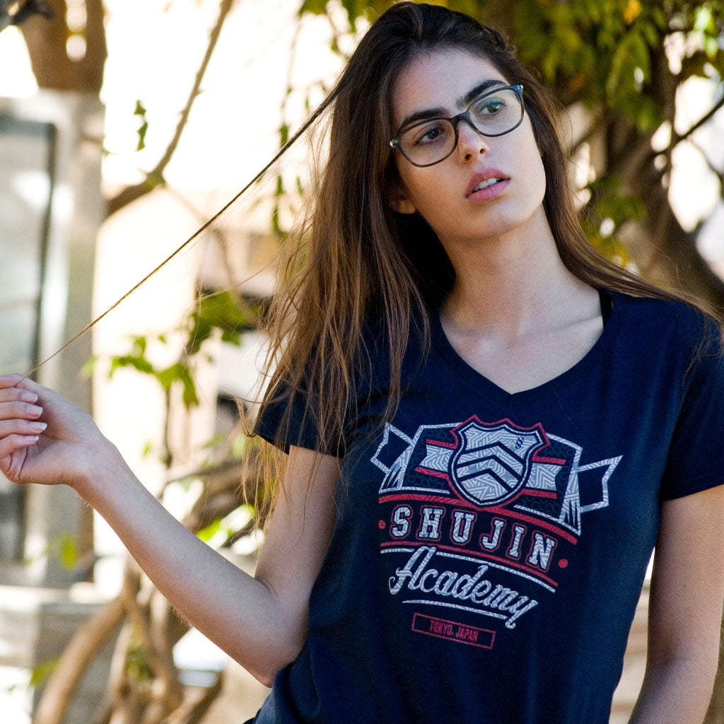 A young woman is wearing the Shujin Academy tee by Eighty Sixed. She is looking off in the distance and twirling her hair standing in front of a blurred background of trees.