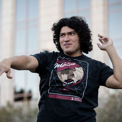 The young man is wearing the Eighty Sixed Persona 5 Welcome to the Metaverse tee. He is making a funny expression while pointing off into the distance with a nice blurred background of buildings.