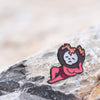 The Shin Megami Tensei V Lazy Miman pin is shown on a rock with some sand with a magnificent blurred background.