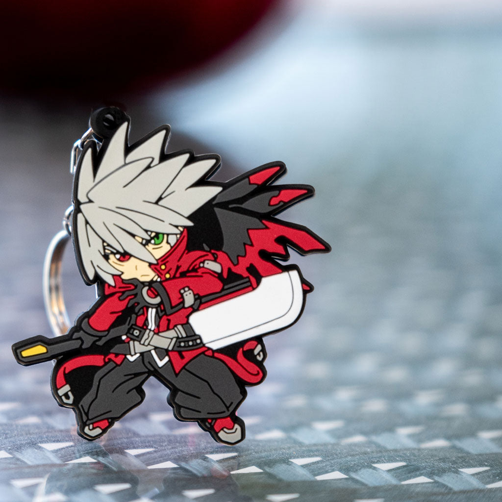 Image featuring the Ragna keychain, a piece of Blazblue merchandise, elegantly positioned on a glass surface with a beautifully blurred background.